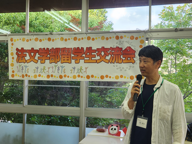 「留学生交流会」のタイトル幕の前で開会の挨拶をする浅田法文学部長